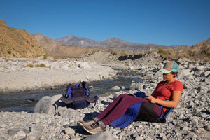 Woman reclining in crazy creek chair next to a river in rocky mountains
