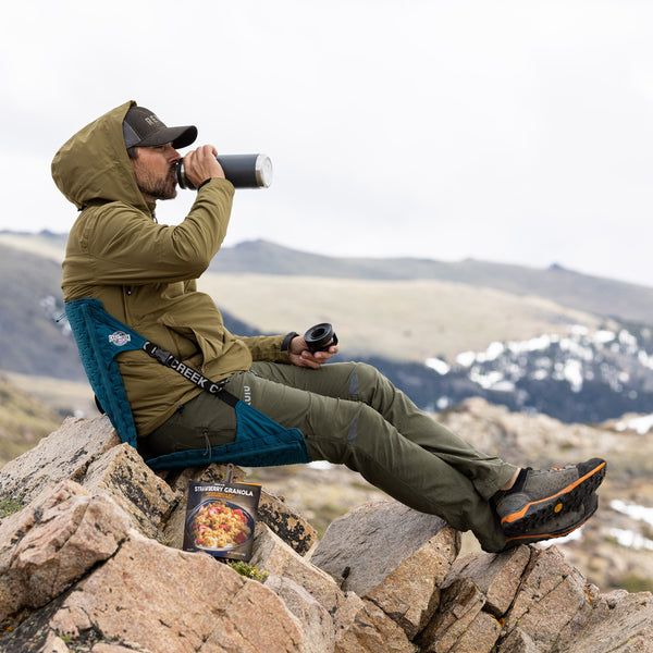 Man drinking water while sitting in a blue crazy creek chair while sitting on the top of a mountain