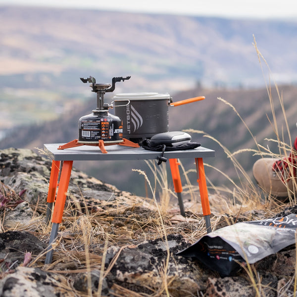 Crazy creek table holding a camping stove and cooking pot in front of mountains