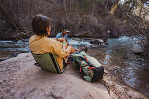 Woman and Man relaxing on the bank of a river with their Osprey x Crazy Creek Talon backpack and Chair
