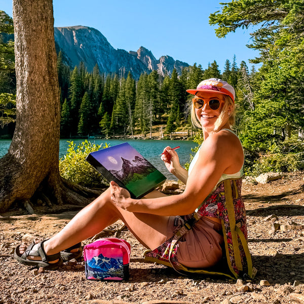 Rachel Pohl Artist painting while sitting in a Crazy Creek chair in front of mountains