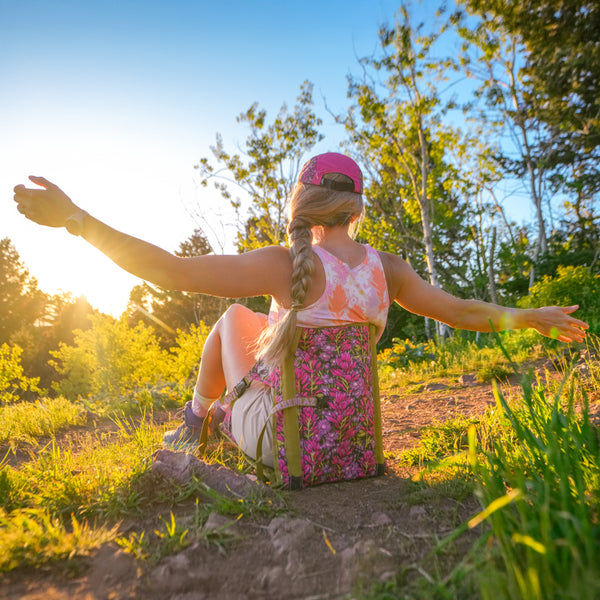 Rachel Pohl sitting in a flowered print crazy creek chair in a forest during sunset with her arms out wearing a pink hat