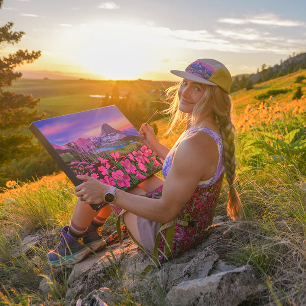 Woman holding a painting of a scenic landscape with flowers in a field while sitting in a Crazy Creek flower printed chair.