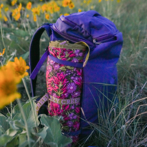 Purple backpack with a backpacking chair rolled up in the water bottle sleeve sitting in a field of yellow flowers