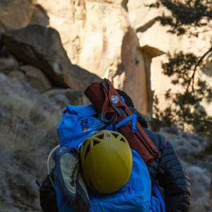 A climber with a pack holding a helmet, climbing shoes, and a rolled up crazy creek chair in the mountains