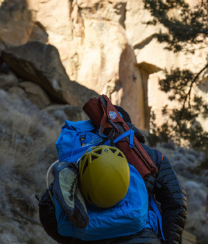 A climber with a pack holding a helmet, climbing shoes, and a rolled up crazy creek chair in the mountains