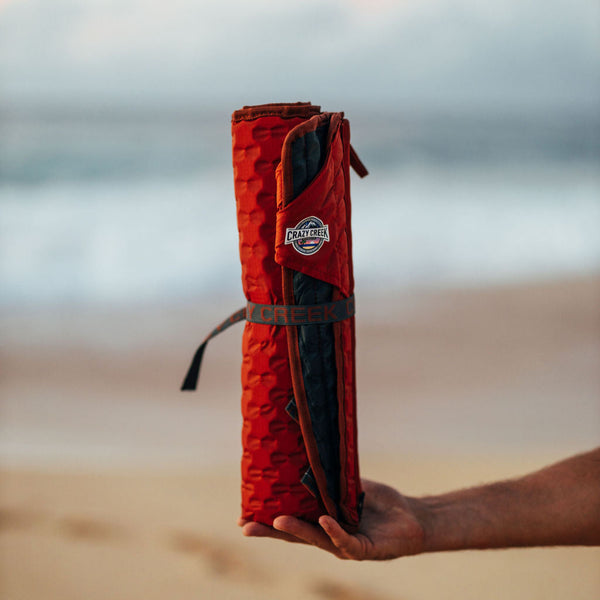 Crazy Creek Hex Chair rolled up and secured with strap - Man holding it in front of a beach
