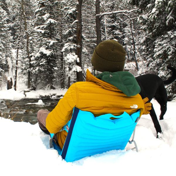 Person sitting in a blue inflatable chair with his dog next to a winter river in the snow