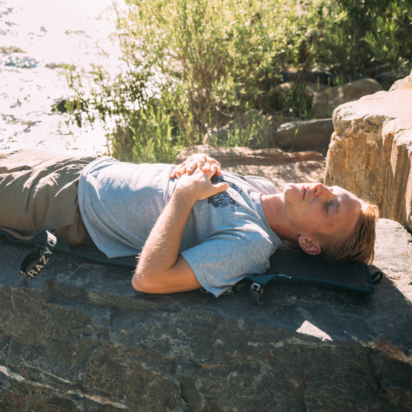 Man lying on a sleeping mat by a body of water with trees in the background