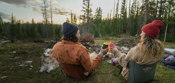 Two people cheersing their drinks while sitting in crazy creek chairs in front of a bonfire next to their dog