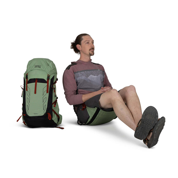 Backpacking man sitting in the Osprey and Crazy Creek Collab Hex chairon white - with backpack sitting next to him